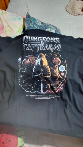Uma camiseta preta é mostrada, pendurada em um fundo desfocado. A camiseta tem um design gráfico na frente. O design apresenta vários capivaras em diferentes poses, com o texto “Dungeons & Capybaras” em letras grandes e estilizadas acima deles. Abaixo da imagem dos capivaras, há um texto menor que diz “Join a fearless group of capybaras on an epic quest for glory, facing fearsome beasts and ancient mysteries. Their adventure begins now.” O design também inclui elementos como um dado e outros detalhes decorativos.

“Dungeons & Capybaras”
“Join a fearless group of capybaras on an epic quest for glory, facing fearsome beasts and ancient mysteries. Their adventure begins now.”