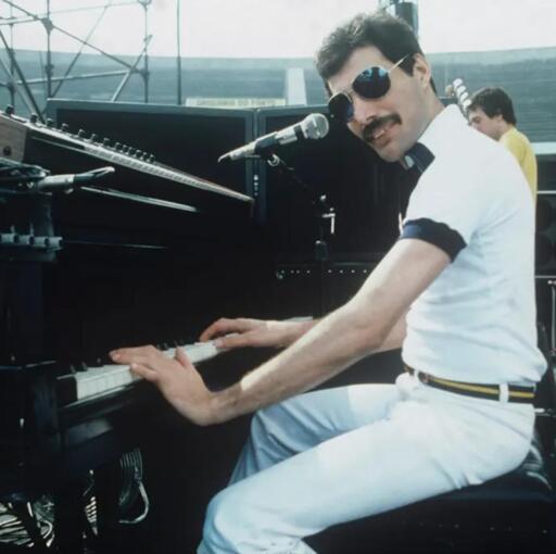 Uma fotografia de um homem sentado em frente a um piano. O homem está de perfil, com as mãos no teclado. Ele usa uma camisa branca e calças brancas, com um cinto fino e dourado. O homem tem um bigode grosso e escuro. Atrás dele, à direita, está outro homem, com as costas voltadas para a câmera. O piano é preto e há microfones posicionados acima dele. O fundo é desfocado, mostrando uma estrutura metálica.